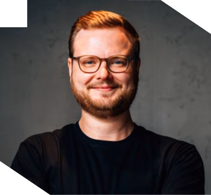 Smiling man with glasses and a beard wearing a black shirt against a gray background.