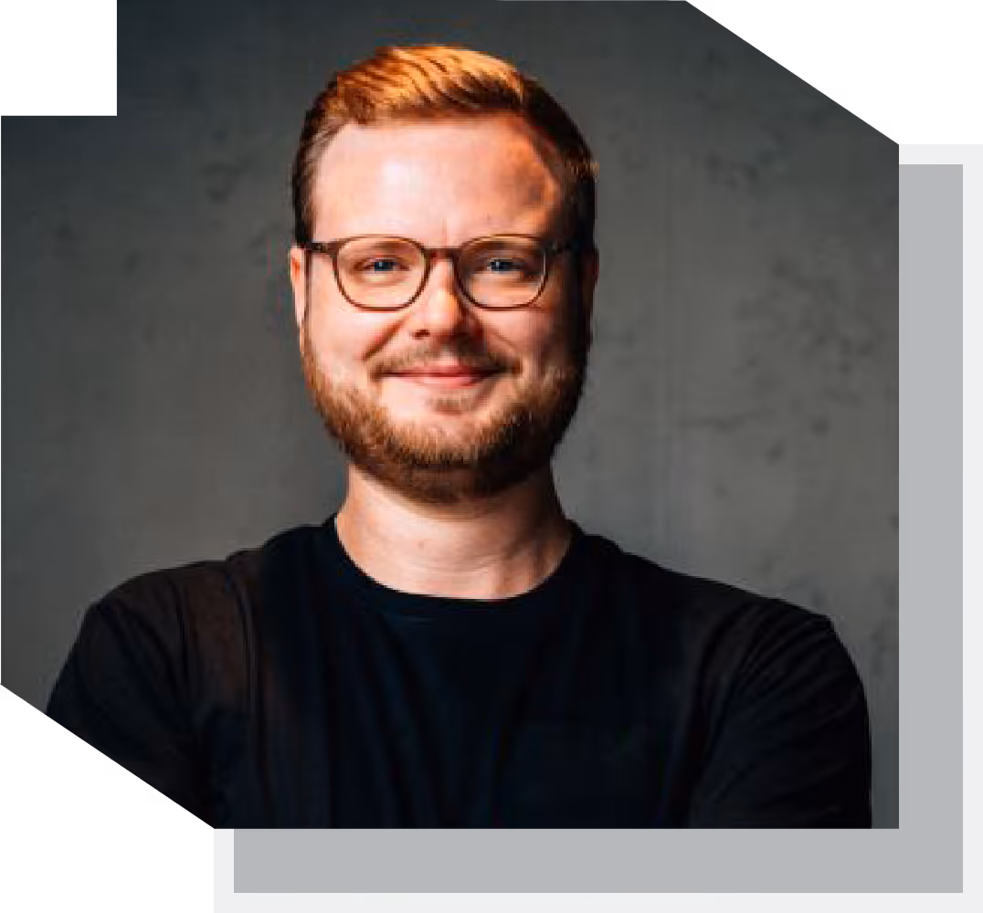 Smiling man with glasses, beard, and short hair wearing a black shirt against a gray background.